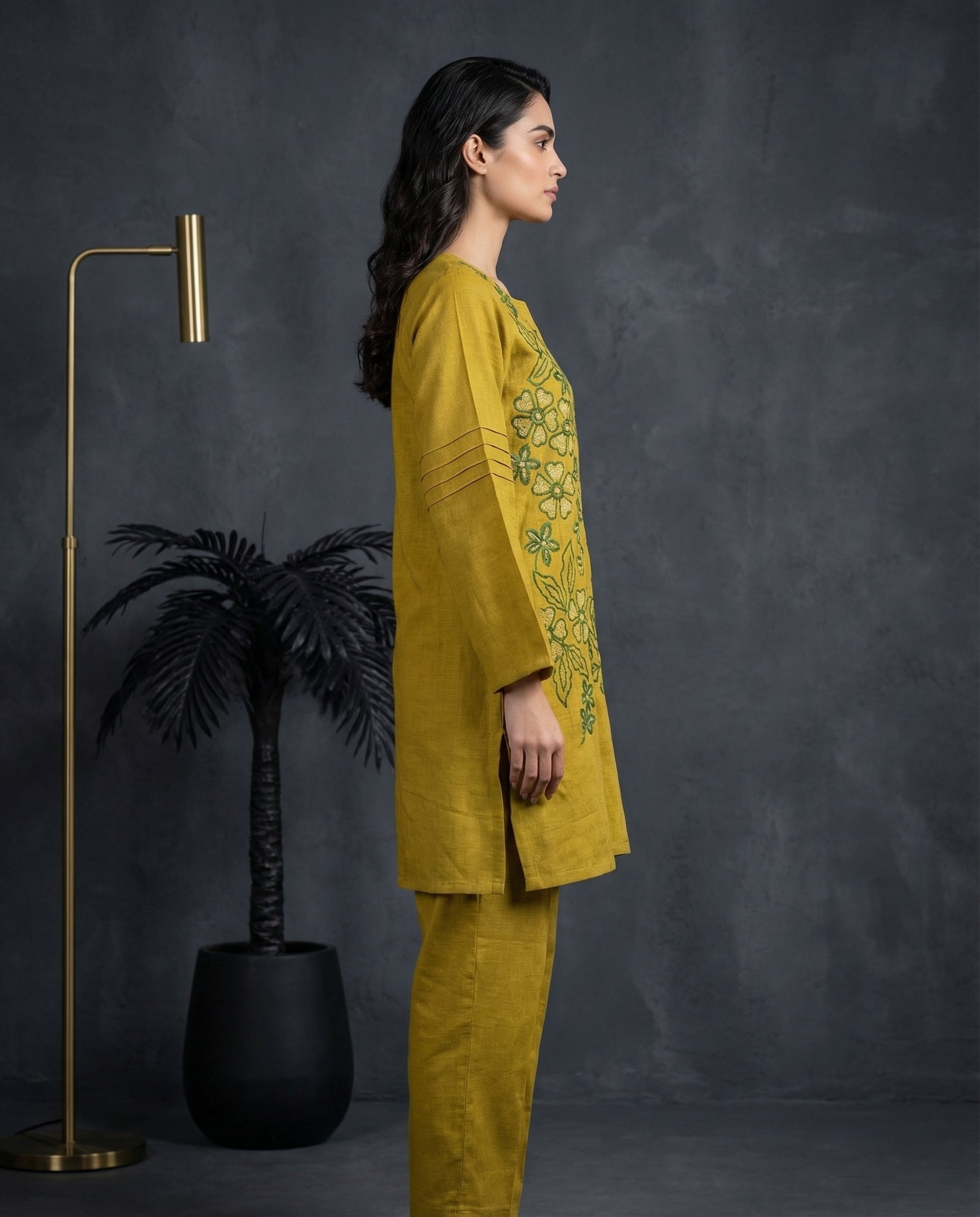 Woman in a mustard yellow outfit with floral embroidery standing against a dark gray background.